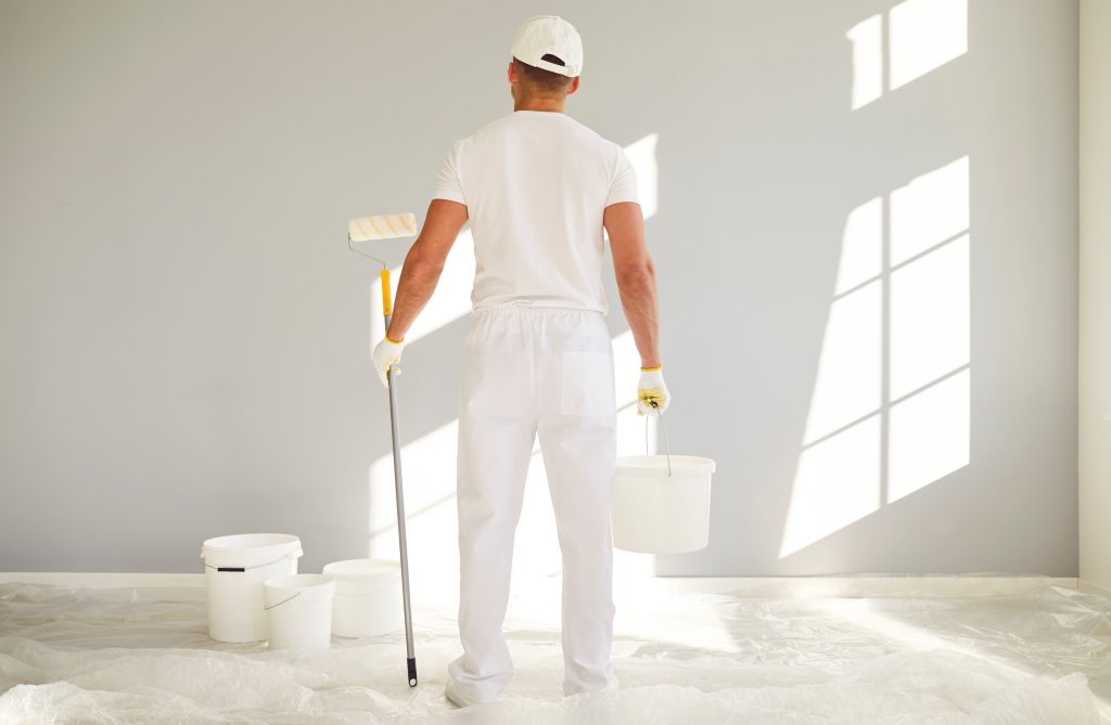 painter holding a bucket and paint roller going painting a grey wall in empty room.